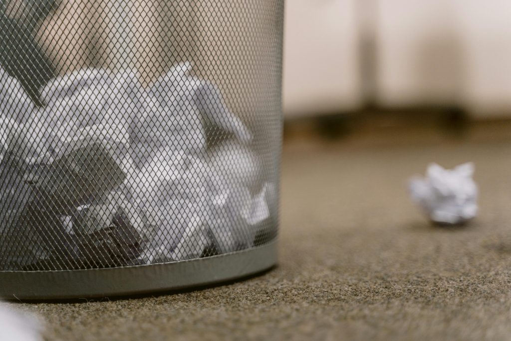 Close-up image of a mesh wire garbage can with scattered crumpled papers on a carpeted floor.